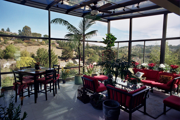 Interior of a sunroom with glass walls and ceiling and a ceiling fan, red couches and armchairs, a dining room table and chairs, and tropical plants inside and a mountainous terrain outside.